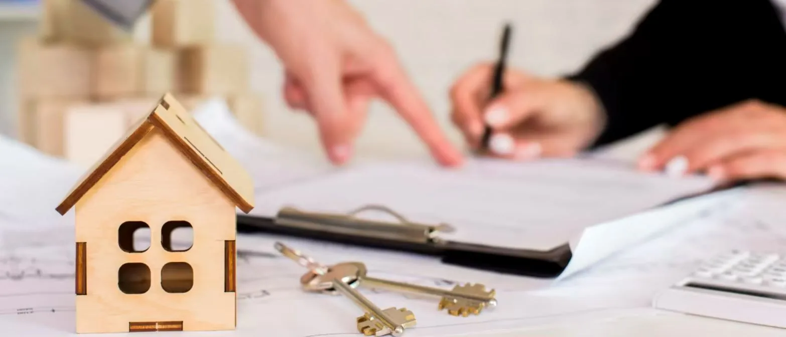 dos personas firmando documentos en una mesa con una maqueta de una casa de madera y dos llaves en primer plano